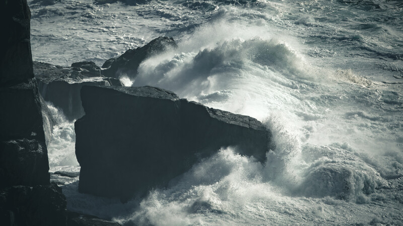 Massive waves crash against dark rocky outcrops with explosive white spray and mist.