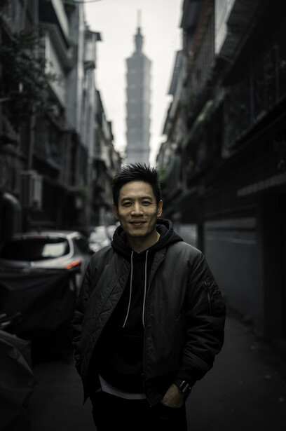 Man in black hoodie standing centered on an urban street with tall buildings framing the composition.