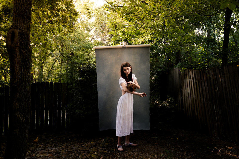 Woman in white dress standing against large gray rectangular panel in wooded setting.