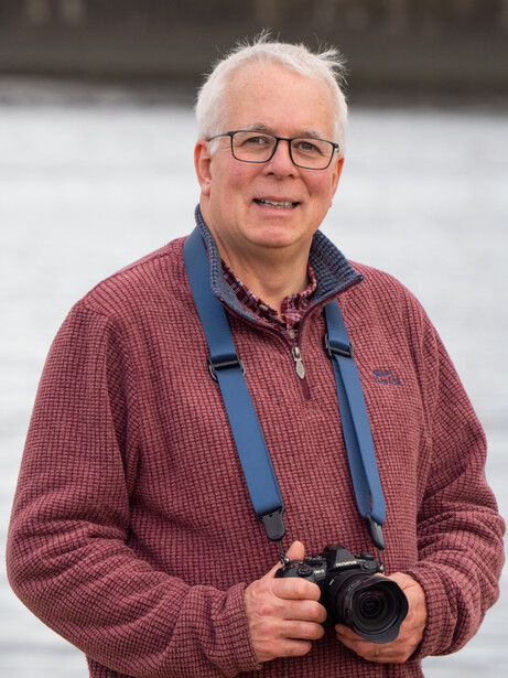Older man with white hair and glasses holding a professional camera with a blue neck strap.