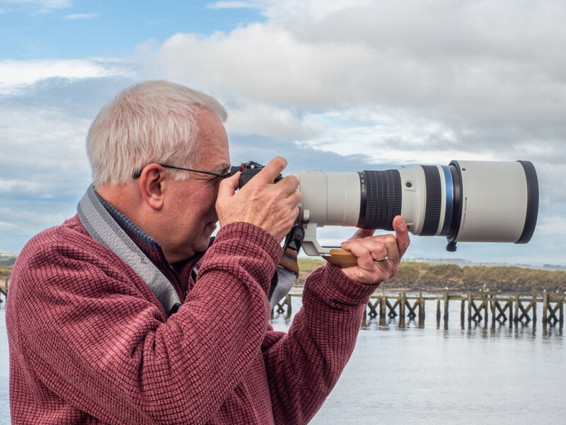 Older photographer with white hair holding a telephoto lens camera to his face, shooting outdoors.