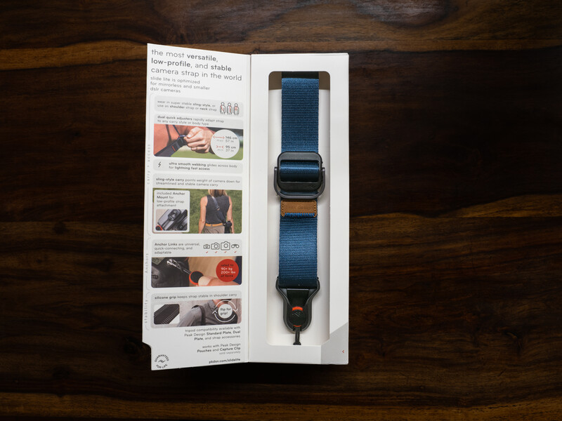Blue camera strap with brown leather accents displayed on packaging against dark wood surface.