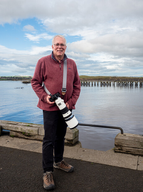 Photographer holding a telephoto lens while standing at a waterside location with wooden pilings visible behind him.