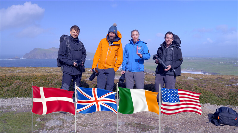 Four hikers holding four flags on a windswept hilltop with coastline visible behind them.
