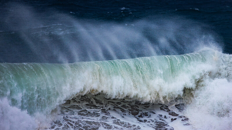 Large ocean wave with white foam crest and turquoise face backlit by dramatic sun rays.