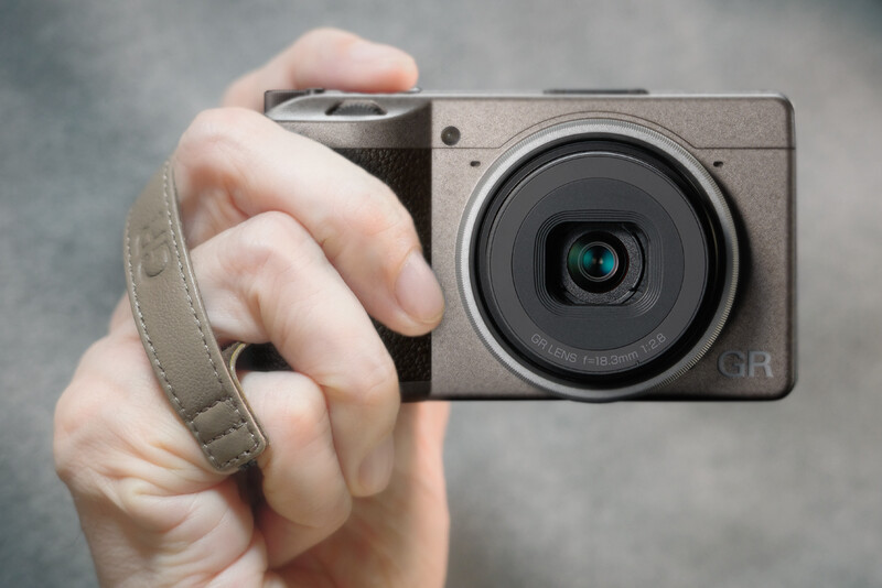 Hand holding a compact digital camera with a large prime lens and fabric wrist strap against a neutral gray background.