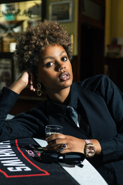 Woman in dark blazer posing at indoor table with hand near face.
