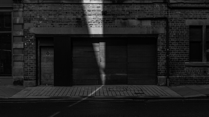 Sunlight streams through a gap between brick buildings, illuminating a storefront with dark metal garage doors.