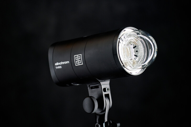 Elinchrom studio flash with reflector mounted on stand against black background.