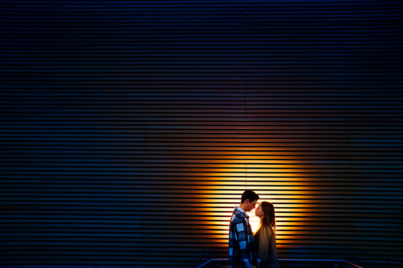 Couple kissing in front of a glowing neon wall with horizontal blue and orange lines.