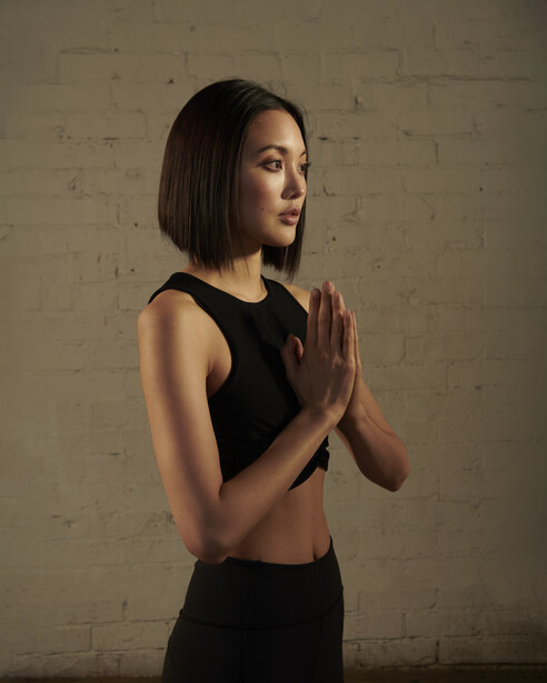 Woman in black sleeveless top with hands pressed together in meditation pose against textured wall.