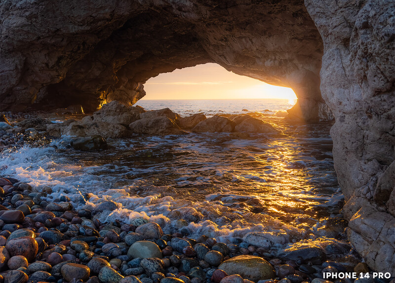 Sea cave opening framing golden sunset light over rocky beach and waves.