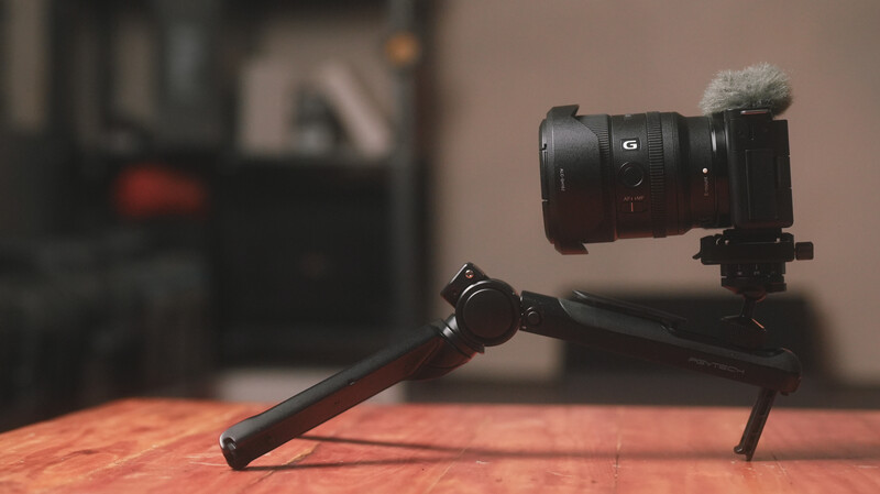 Sony mirrorless camera with microphone mounted on tripod with wooden base against blurred interior background.