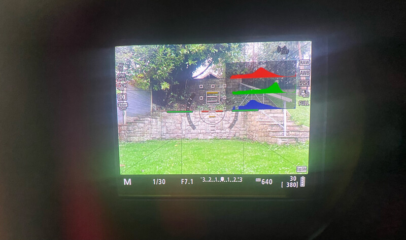 Camera viewfinder display showing a residential property with house and outbuilding framed by a stone wall and green lawn.