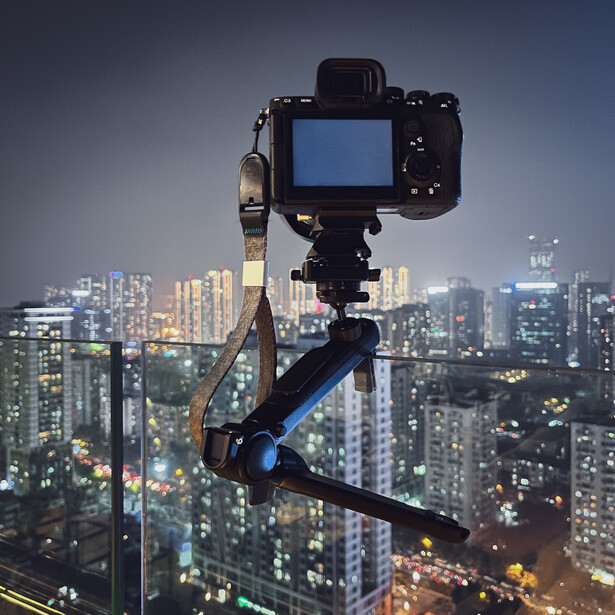 Camera on tripod with articulating arm mounted on high-rise building overlooking illuminated city skyline at dusk.