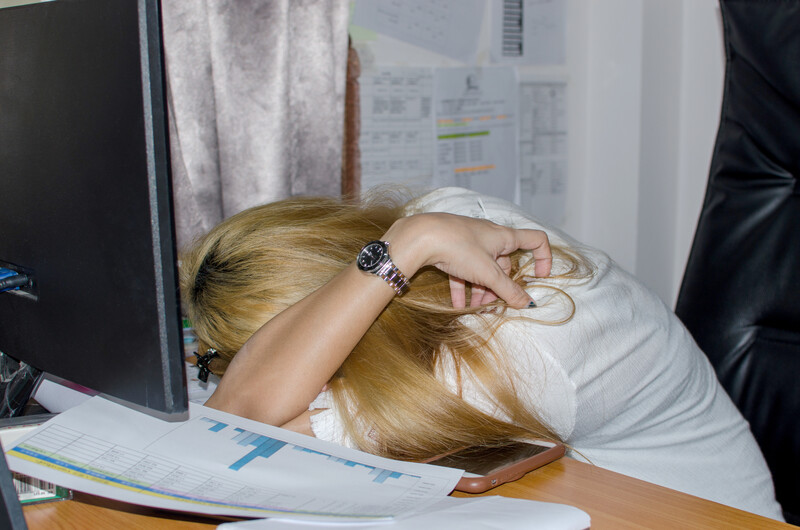 Woman resting her head on a desk while working at a computer with papers and documents nearby.