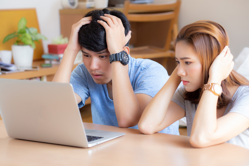 Two young adults in blue shirts looking stressed while viewing a laptop screen together at a wooden desk.