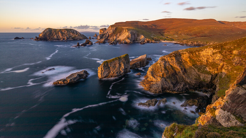 Rocky sea stacks and cliffs glowing in golden hour light, with smooth water and distant islands under a twilight sky.