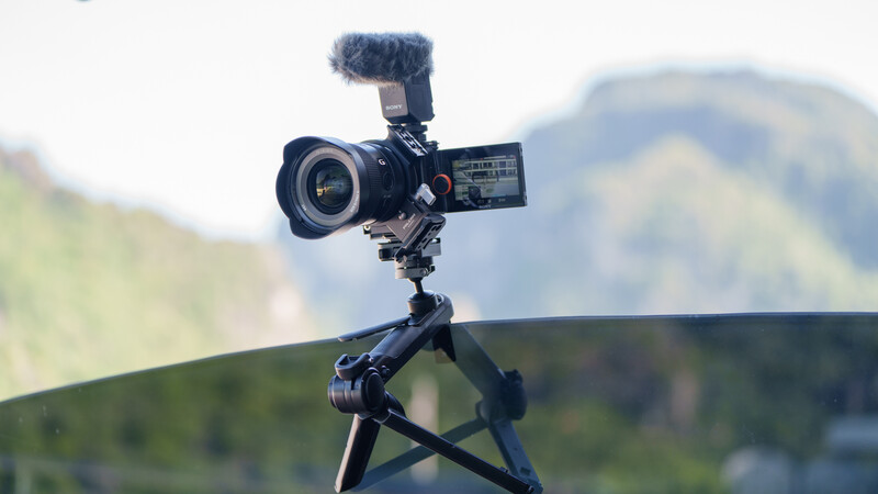 Compact cinema camera mounted on tripod with articulating monitor, external microphone, and telephoto lens against mountainous backdrop.