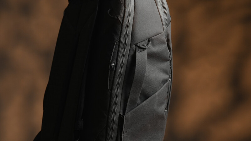 Close-up detail of a black and gray technical backpack showing zippered pockets and textured fabric panels.