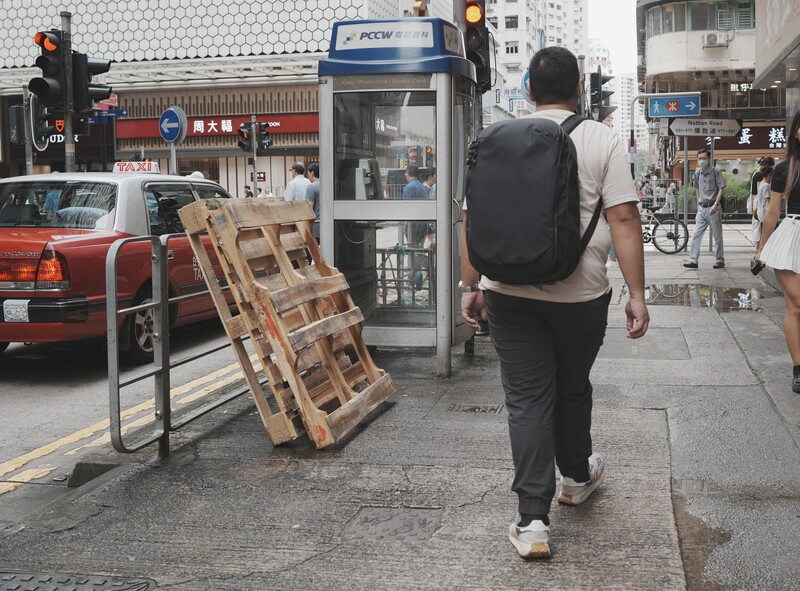 Street scene with person wearing backpack jogging past wooden pallets and storefronts.