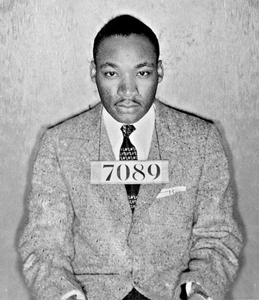 Black and white mugshot portrait of a man wearing a suit and tie, holding a numbered placard reading 7089.