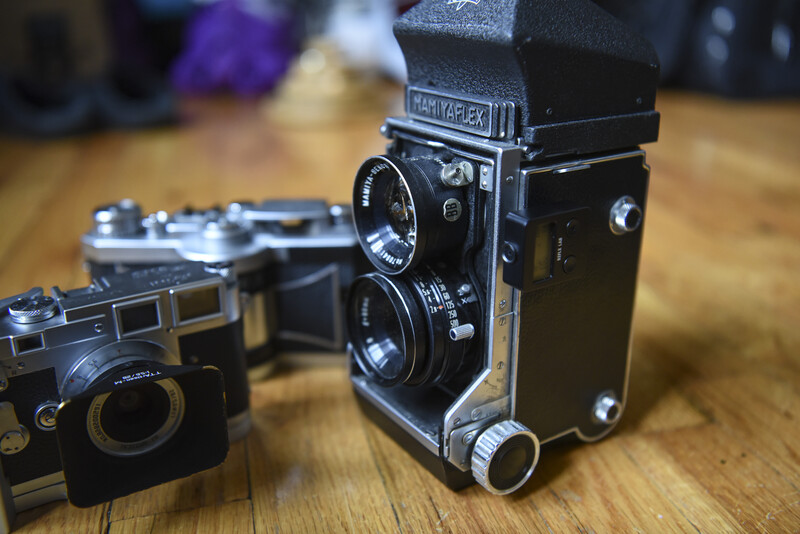 Collection of vintage film cameras arranged on a wooden surface, including a twin-lens reflex camera with flash attachment in sharp focus.
