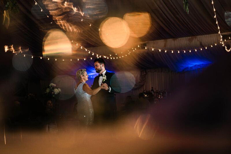 Couple dancing during first dance with dramatic dry ice fog and warm string lights overhead.