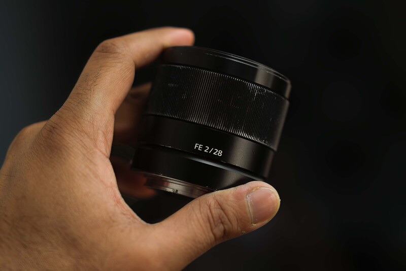 Hand holding a compact camera lens with FE 2/28 markings against a dark background.