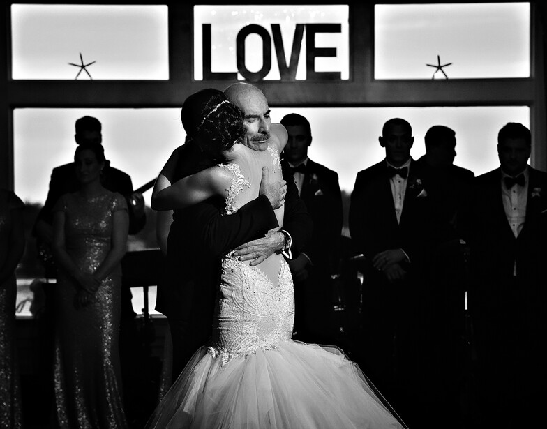 Bride in beaded strapless gown stands center frame surrounded by silhouetted wedding guests, with illuminated "LOVE" sign above.