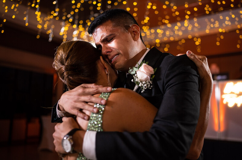Groom in black suit embracing bride in white wedding dress during first dance under warm golden bokeh lights.
