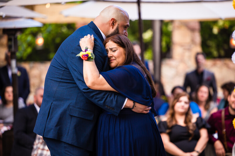 A couple in navy blue formal attire embrace during an outdoor ceremony, with guests visible in the soft-focused background.