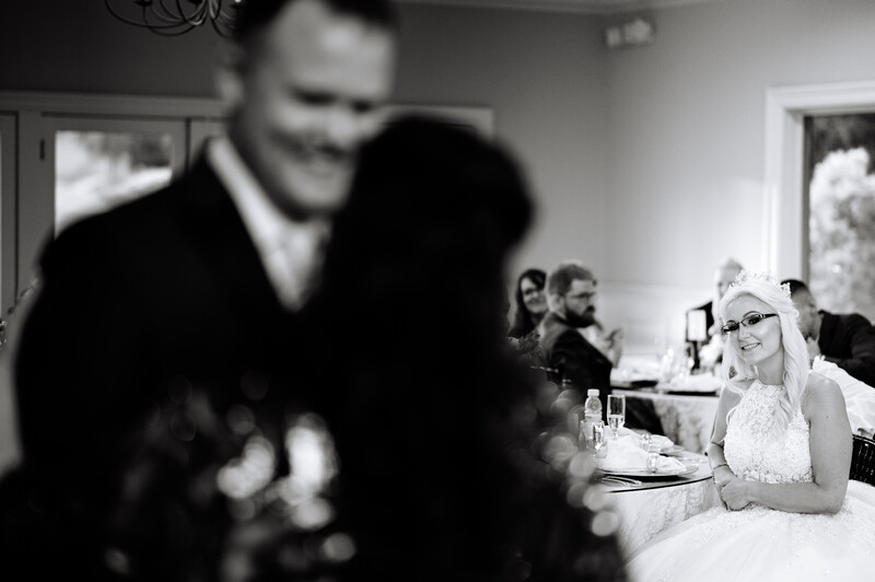 A bride in a white lace dress sits at a reception table, focused and smiling, while blurred guests and a couple embrace in the background.