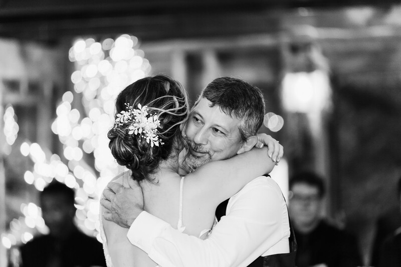 Bride and groom embracing during first dance, shot in black and white with soft bokeh lights in background.