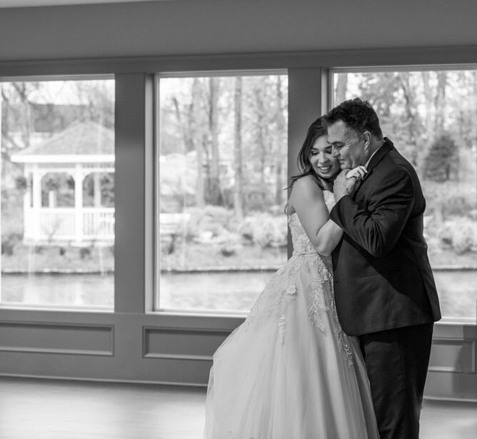 Groom embracing bride from behind while both look at camera in black and white wedding portrait.