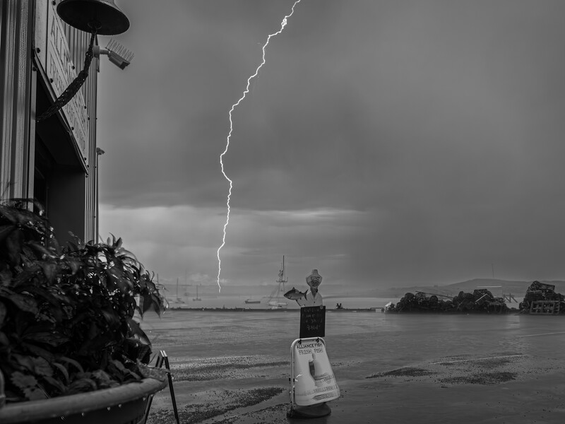 Lightning bolt striking across stormy sky above a waterfront with boats and industrial structures visible on the shore.