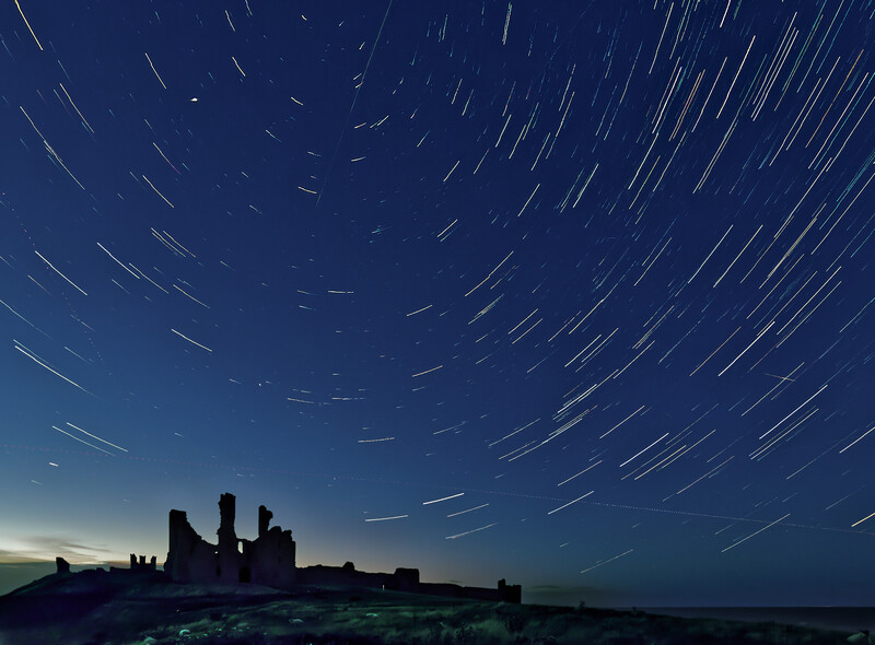 Silhouetted castle ruins against a night sky filled with star trails in concentric circular patterns.