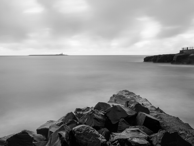 Rocky breakwater extends into a misty seascape with a distant lighthouse under overcast skies.