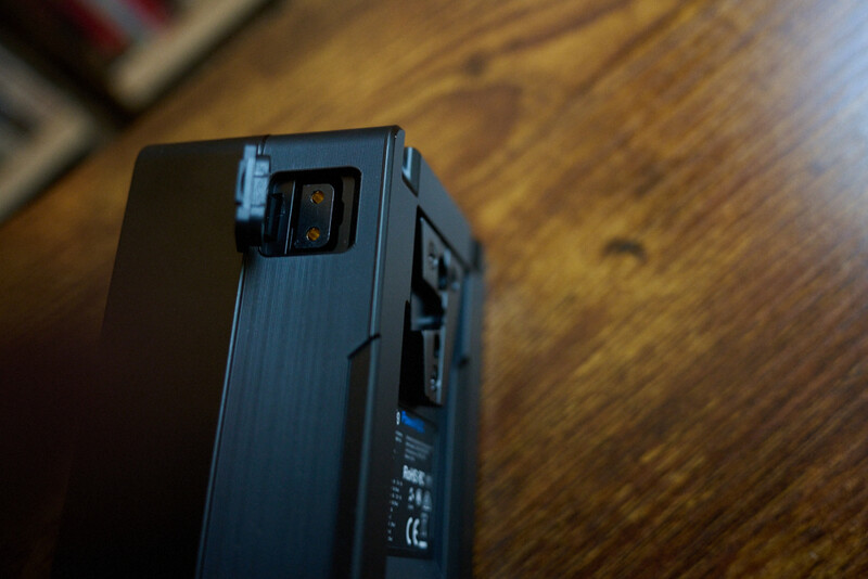 A compact electronic device with a dark textured surface and blue accents sits on a wooden table, photographed at an angle.