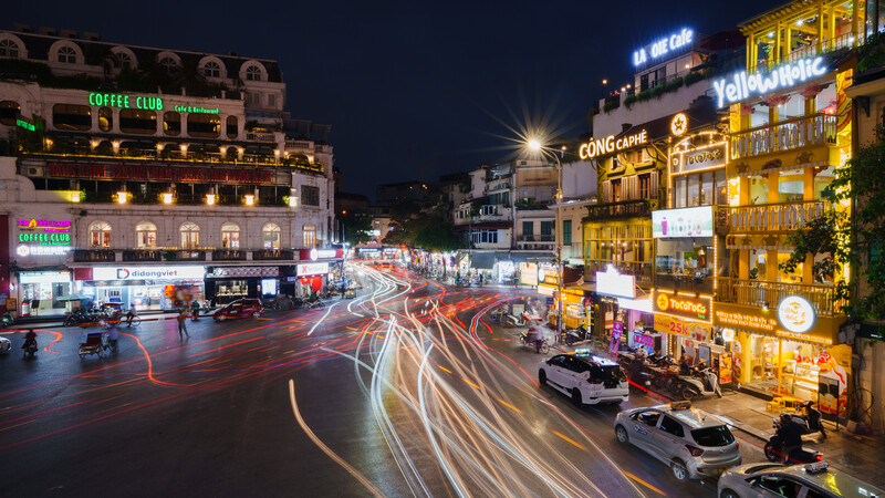Urban intersection at night with light trails from vehicle traffic and illuminated storefronts.