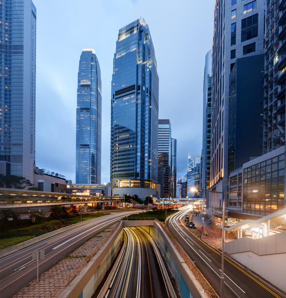 Urban highway with motion-blurred light trails from vehicles passing beneath illuminated skyscrapers at dusk.