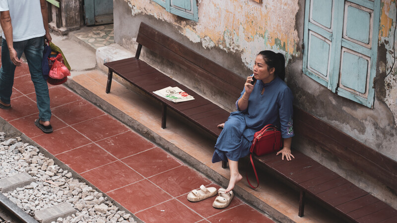 Woman sitting on a wooden bench against a weathered building wall, eating from a plate.