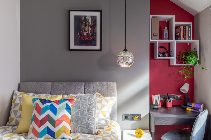 Modern bedroom featuring gray walls, colorful patterned pillows on bed, and hot pink shelving unit with books and plants.