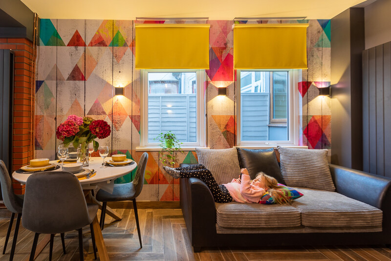 Modern apartment interior with geometric wall art, person relaxing on sofa, and bright yellow window treatments.