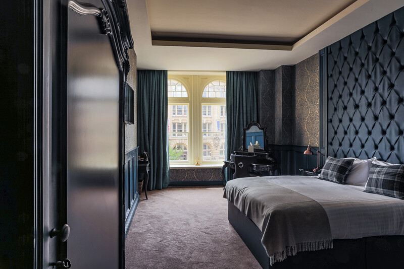 Luxury hotel bedroom with quilted headboard, modern furnishings, and large arched windows overlooking a city street.
