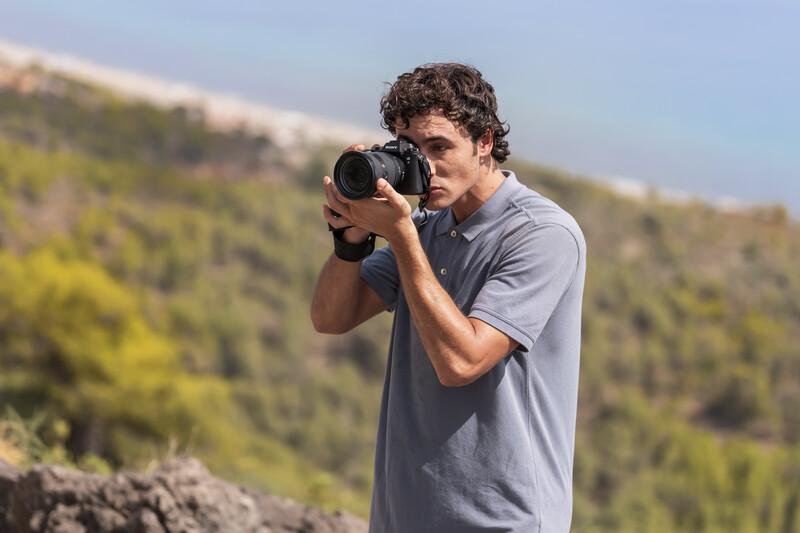 Photographer holding a telephoto lens to his eye while composing a shot outdoors.