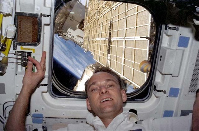 Astronaut inside a spacecraft window looking out at solar panels and Earth's surface below.