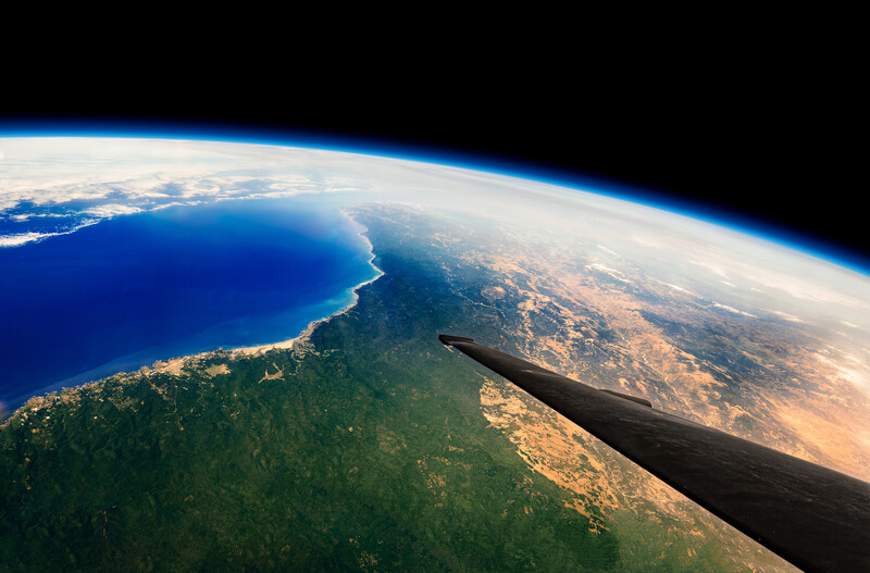 Aerial view from aircraft window showing curved Earth horizon, coastline, ocean, and forested landmass below.