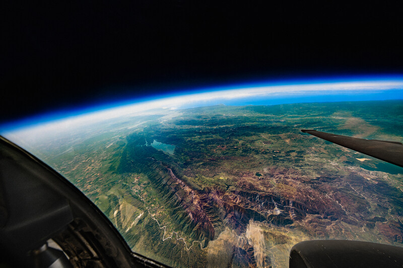 Earth's curved horizon and atmospheric layers viewed from an aircraft window, showing patchwork terrain and blue ocean below.