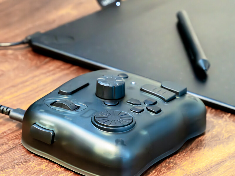 Black audio interface with knobs, faders, and buttons on a wooden desk beside a tablet and stylus.
